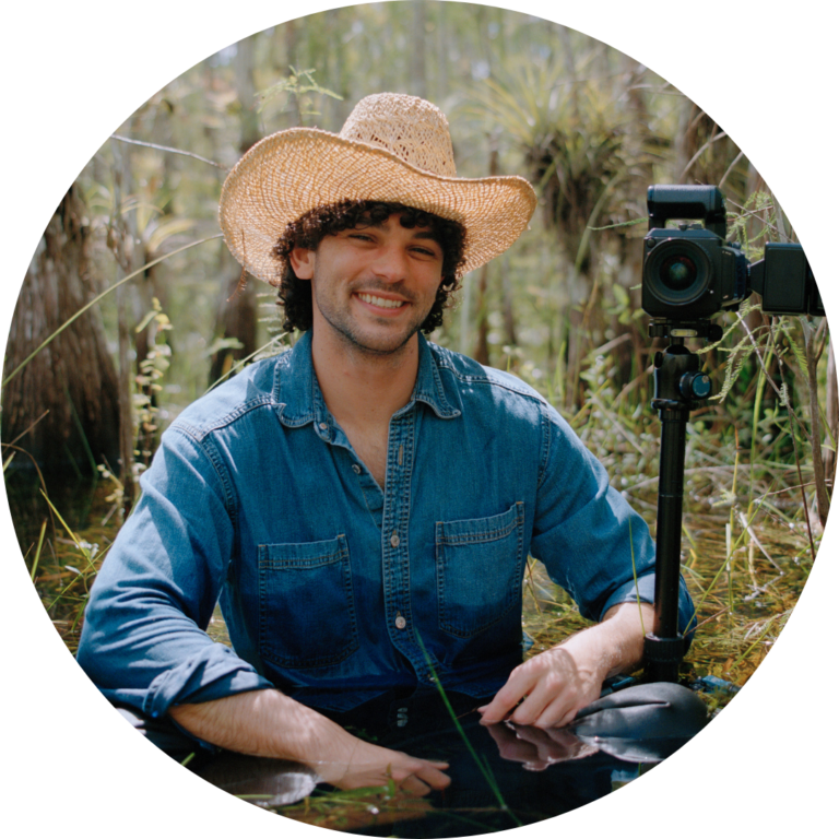 A person in a denim shirt and straw hat sits in shallow water outdoors, smiling at the camera, with a camera on a tripod beside them among tall grasses—capturing the creative spirit inspired by the Vizcaya Contemporary Arts Program.