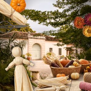 A table with corn husk dolls, mini pumpkins, dried corn, and flowers in front of a Vizcaya Village.