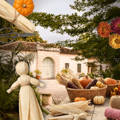 A table with corn husk dolls, mini pumpkins, dried corn, and flowers in front of a Vizcaya Village.