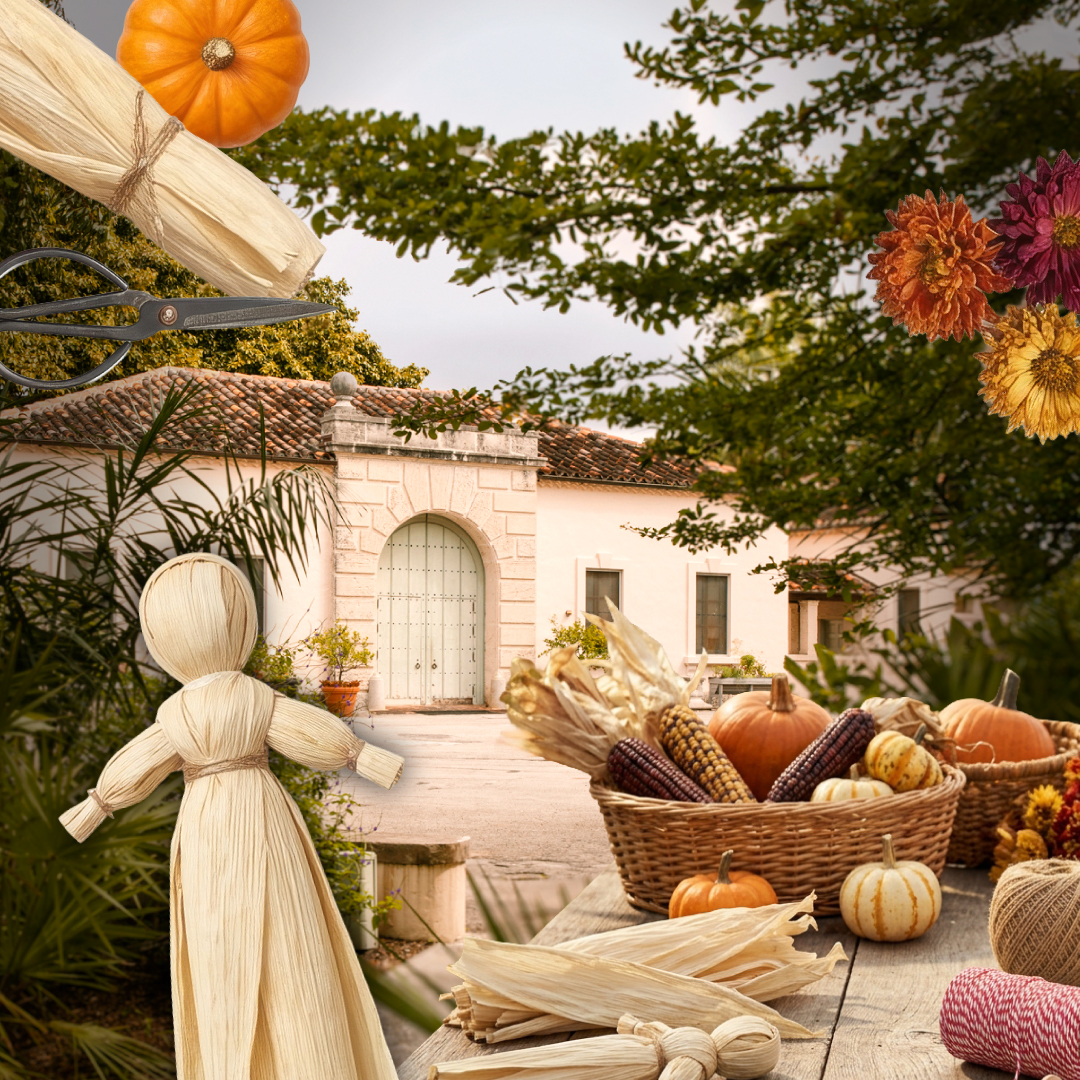 A table with corn husk dolls, mini pumpkins, dried corn, and flowers in front of a Vizcaya Village.