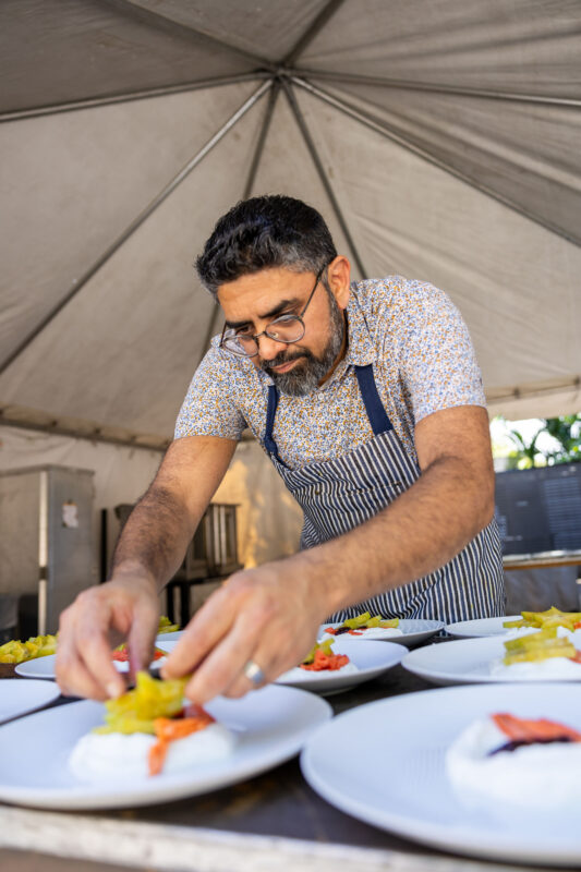 A chef wearing glasses and a striped apron plates white dishes topped with yellow fruit and red garnish.