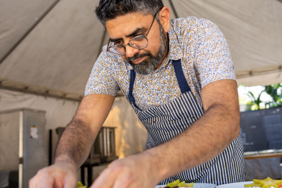 A chef wearing glasses and a striped apron plates white dishes topped with yellow fruit and red garnish.