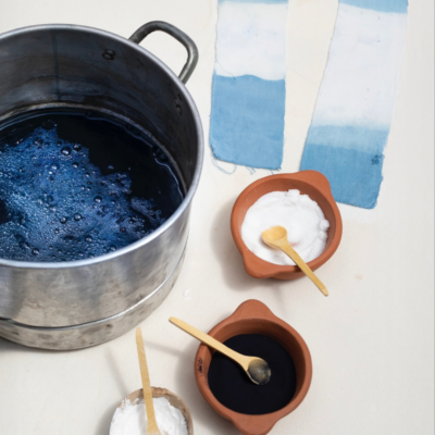 A metal pot filled with dark blue indigo dye next to bowls of powder and dye samples on fabric strips.