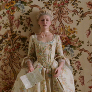 A woman in an 18th-century gown and choker stands in front of floral wallpaper holding a handwritten letter.